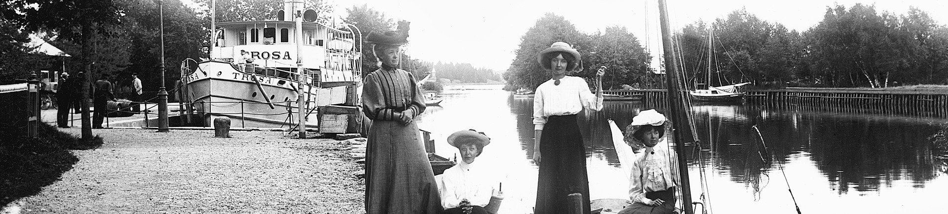 Fyra kvinnor sitter och står vid hamnen i Trosa medan ett ångfartyg står förtöjt bakom dem