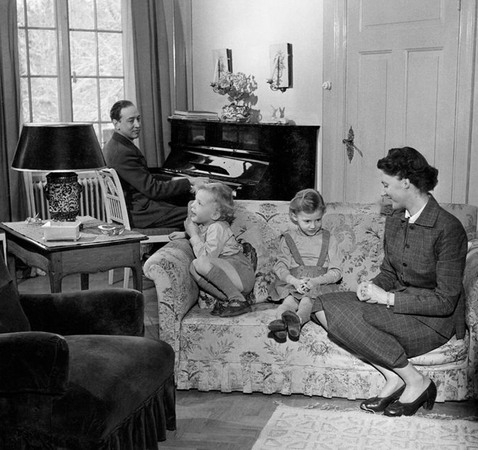 En kvinna sitter med två barn i en blommig soffa. Bakom dem sitter en man vid pianot, framför ett stort fönster.