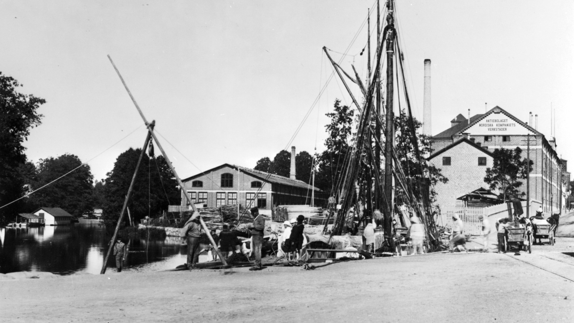 Svart-vitt foto på NK:s möbelfabrik vid Nyköpingsån i början av 1900-talet. Några arbetare syns och segelbåtar ligger vid kajen. 