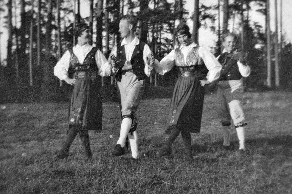 Två kvinnor och två män i Vingåkersdräkter dansar folkdans på en gräsmatta, i bakgrund syns en skog.