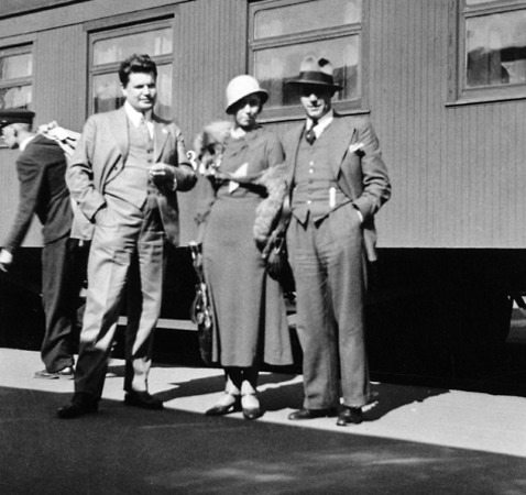 Två män i kostym och en kvinna i lång klänning står framför ett tåg
