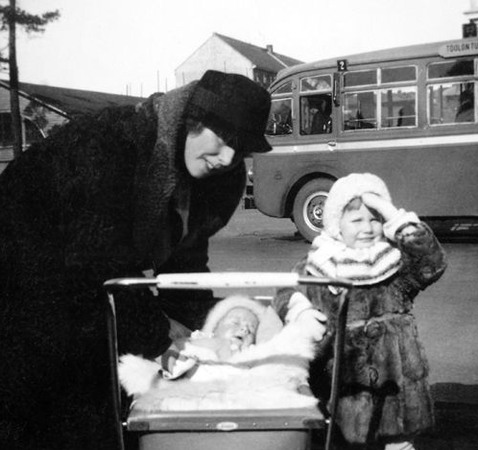 En kvinna i svart kappa och hatt står lutat över en barnvagn med ett sovande spädbarn. På andra sidan om vagnen står en liten flicka i pälsjacka och vit mössa.