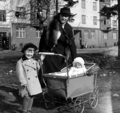 En kvinna i svart kappa och mössa står bakom en vagn med ett litet barn. Framför vagnen står en flicka i ljus kappa och svart hatt. I bakgrunden syns hyreshus och enstaka träd.