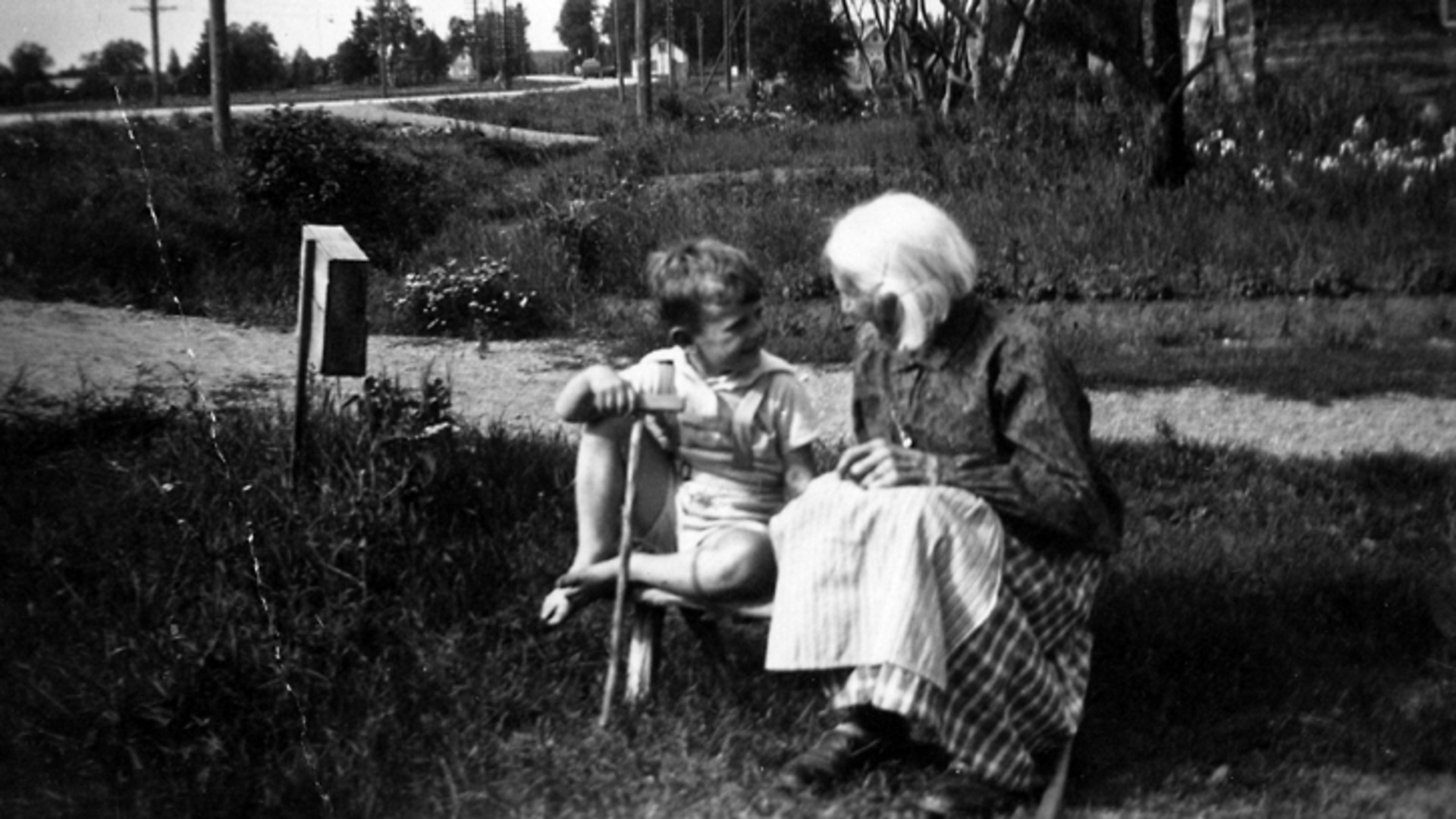En pojke sitter och pratar med en äldre kvinna, på gräsmattan framför ett hus på landet