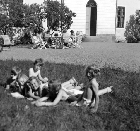 En grupp med barn, i somrig klädsel, ligger i gräset framför ett viss hus.