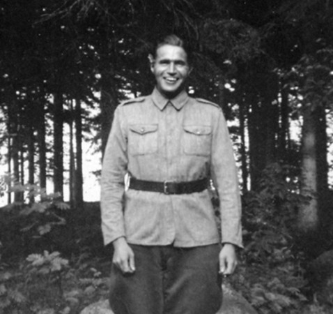 En man i uniform står i ett skogsparti och ler mot kameran