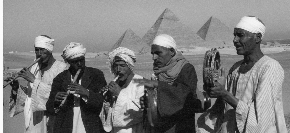 Fem män, iklädda kaftaner och sjalar, med vita huvudbonader spelar flöjt och tamburin. I bakgrunden syns tre pyramider på avstånd.