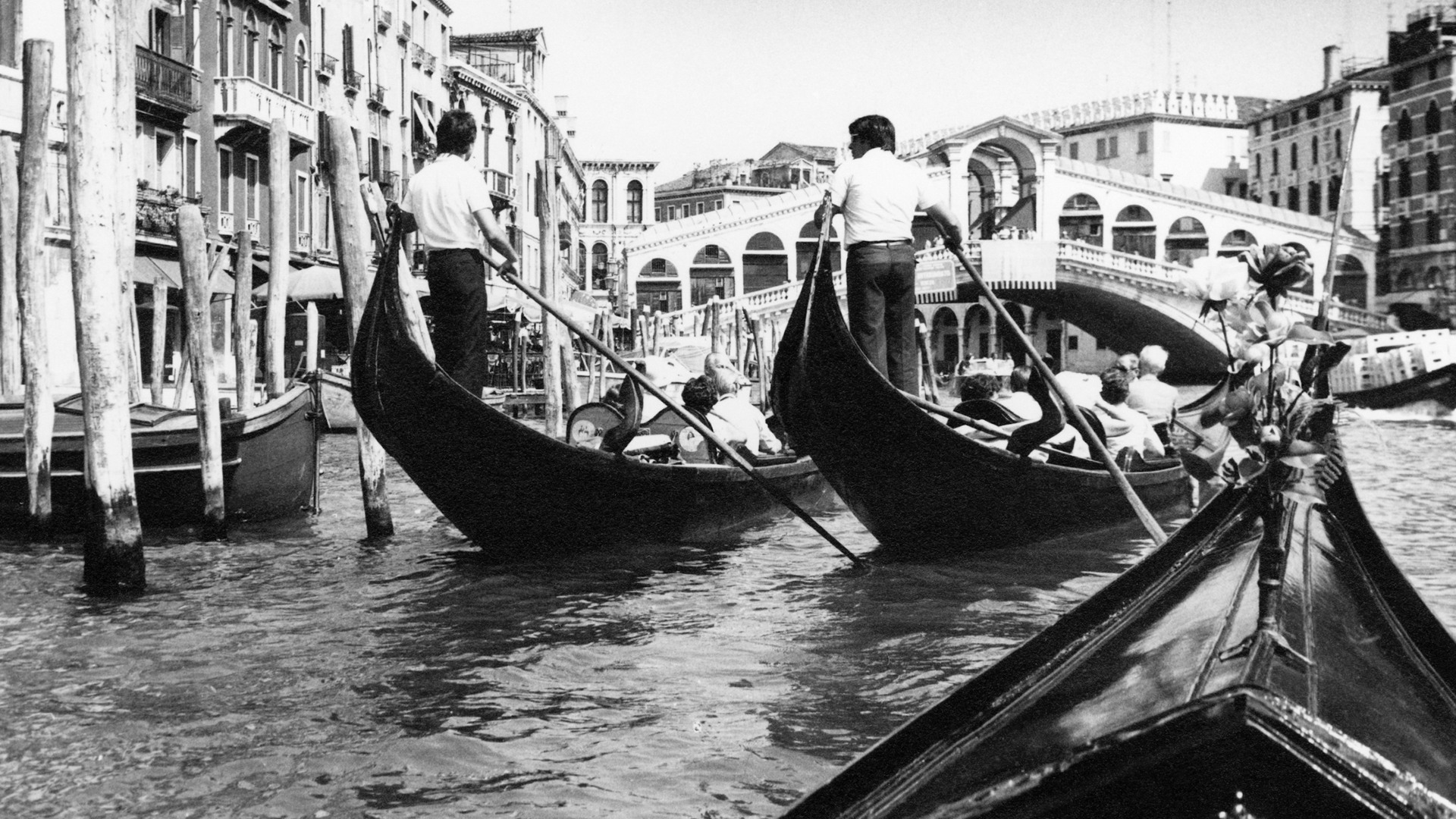 Två män står i aktern på varsin båt och paddlar längsmed en flod som kantas av en stadsmiljö. I båtarna sitter ett par stycken människor.