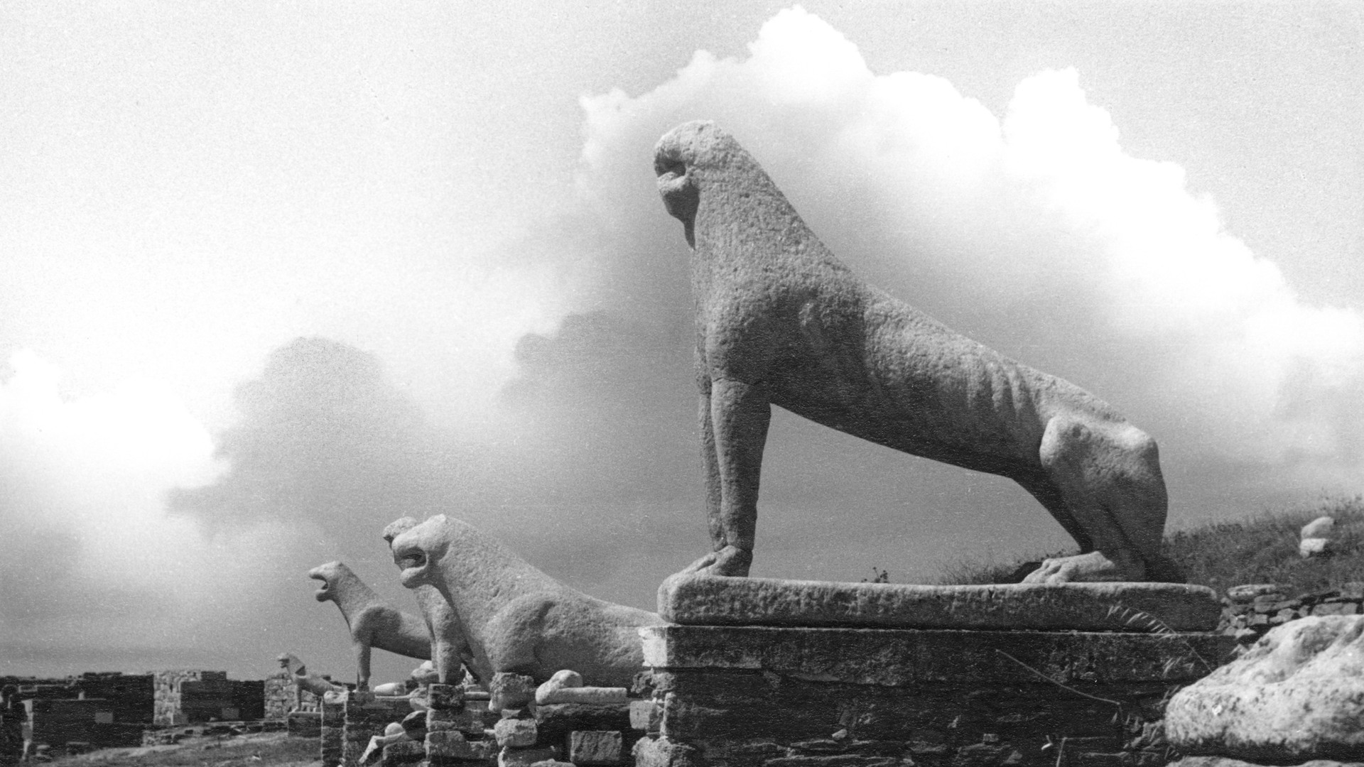 En rad med sten-statyer, föreställande något slags fyrbent djur, sträcker sig mot horisonten. Moln tornar upp sig över himlen. Omgivningen är karg.