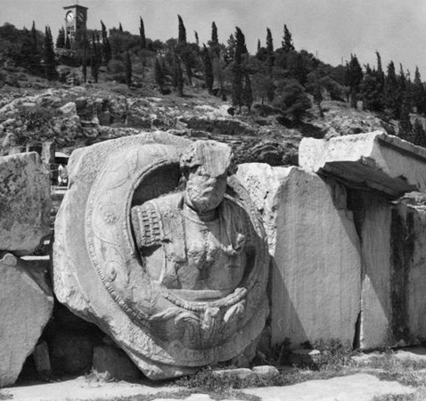 En sliten skulptur av en pampig herre står lutad mot en stenkant. I bakgrunden reser sig en kulle med vindpinade enar och högst uppe på toppen kan man skymta ett kapell.