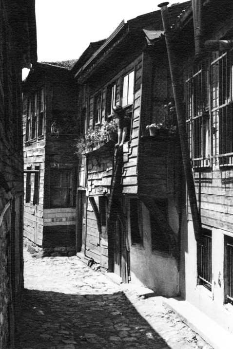 En jättesmal kullerstensgata kantas av låga trähus. I ett fönster står en kvinna och skakar ett tyg.