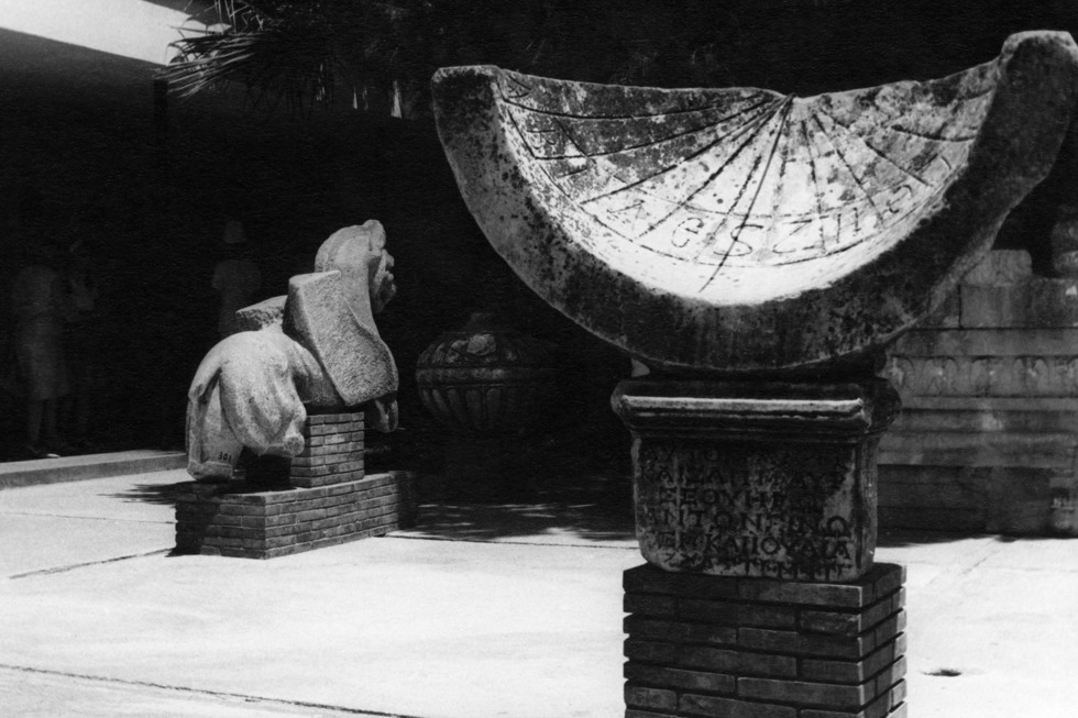 Ett halvcirkelformat objekt i sten, med inskribtioner, i bakgrunden en staty av ett hästliknande djur, bakifrån. 