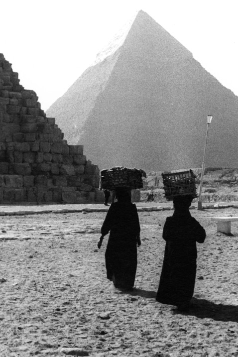 Två personer vandrar, med korgar balanserade på sina huvuden, mot en pyramid. 