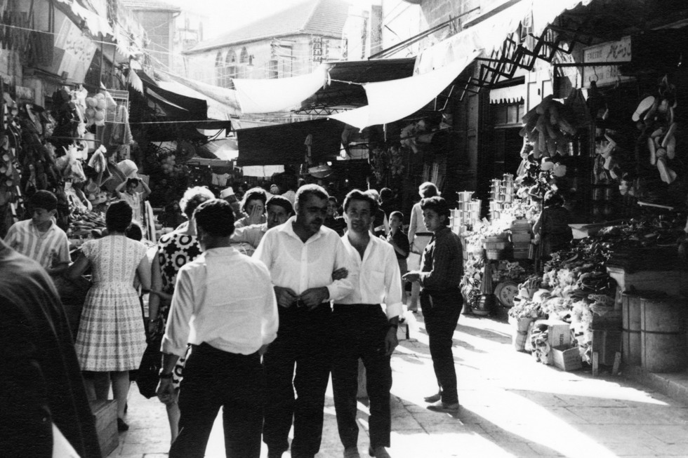 En smal gata, full av människor, kantas av butiker som har ställt ut sina varor på gatan. I mitten av bilden går en grupp män i vita skjortor och svarta kostymbyxor.