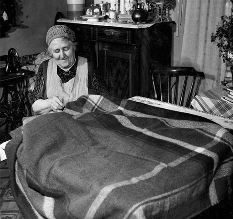 En äldre kvinna sitter med ett stort tyg utbrett över ett bord och syr för hand. 