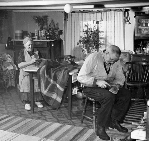 En tant och en farbror sitter i ett litet rum med lågt i tak, linoleum matta och trasmattor på golvet. Kvinnan syr för hand i ett stort tyg, mannen lagar en sko. 