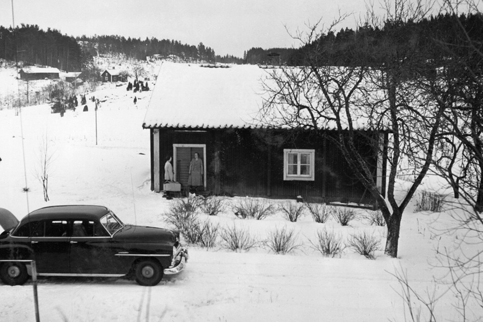 En liten röd stuga i ett snöigt landskap. Framför stugan står en svart bil, i 60-talsstil parkerad. Framför stugan står två personer och samtalar.