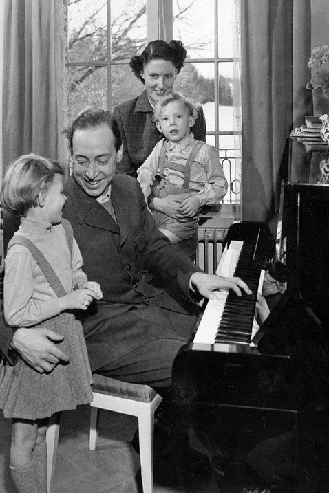 En man sitter vid ett piano och håller om en liten flicka som står bredvid honom. Bakom paret sitter en kvinna med en liten pojke i famnen, framför ett stort fönster. De ser glada ut och den lilla flickan skrattar. 