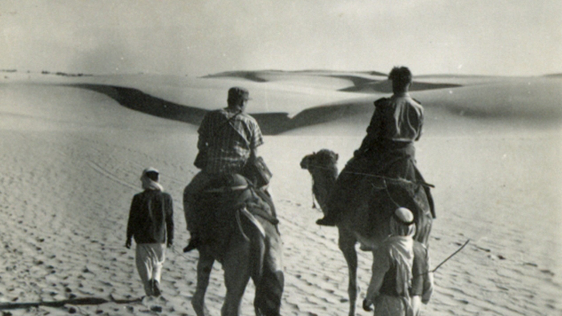 Två män rider på kameler i öknen, en man går framför och en bakom