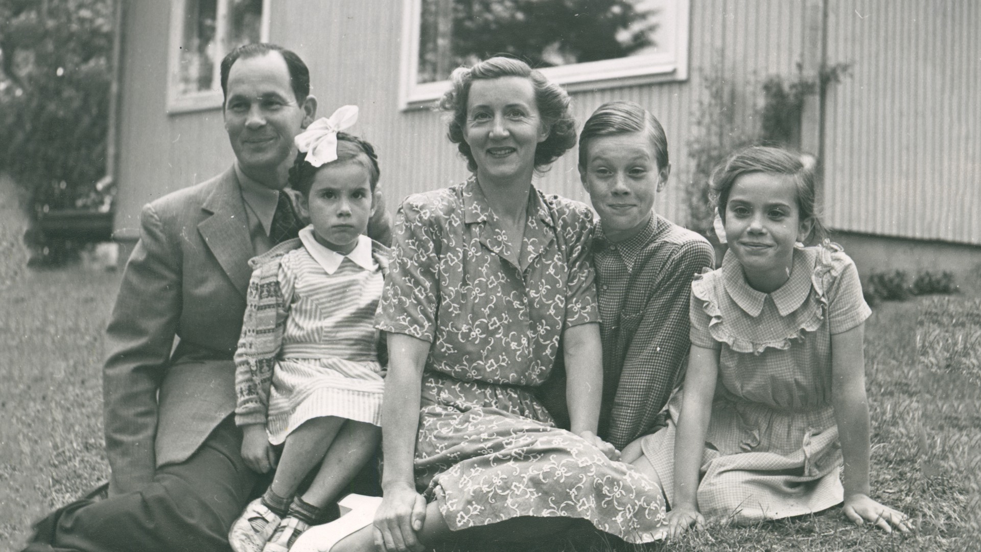 Karin Wohlin sitter i mitten, omgiven av sin familj bestående av man och tre barn. De sitter på gräset framför ett hus.