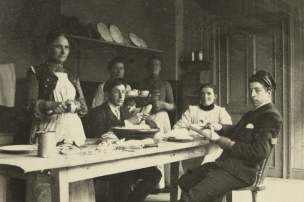 Flera kvinnor och män jobbar i ett kök vid ett bord och skalar grönsaker. 