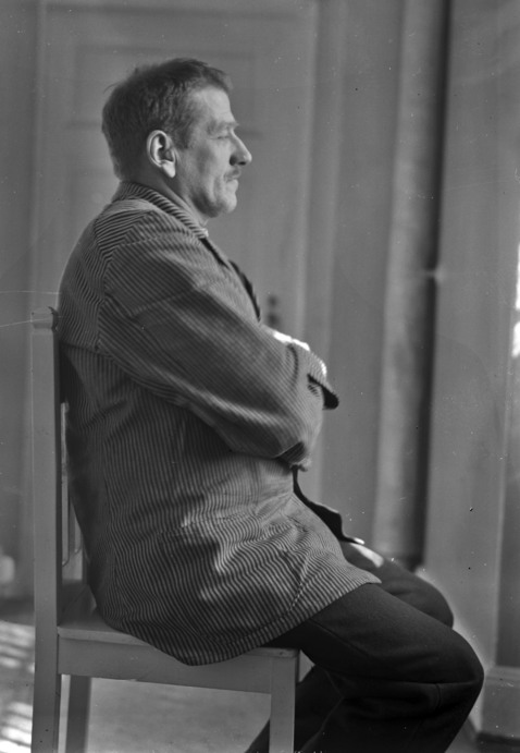 Manlig patient i profil, sitter på stol och tittar ut genom ett fönster med armarna i kors