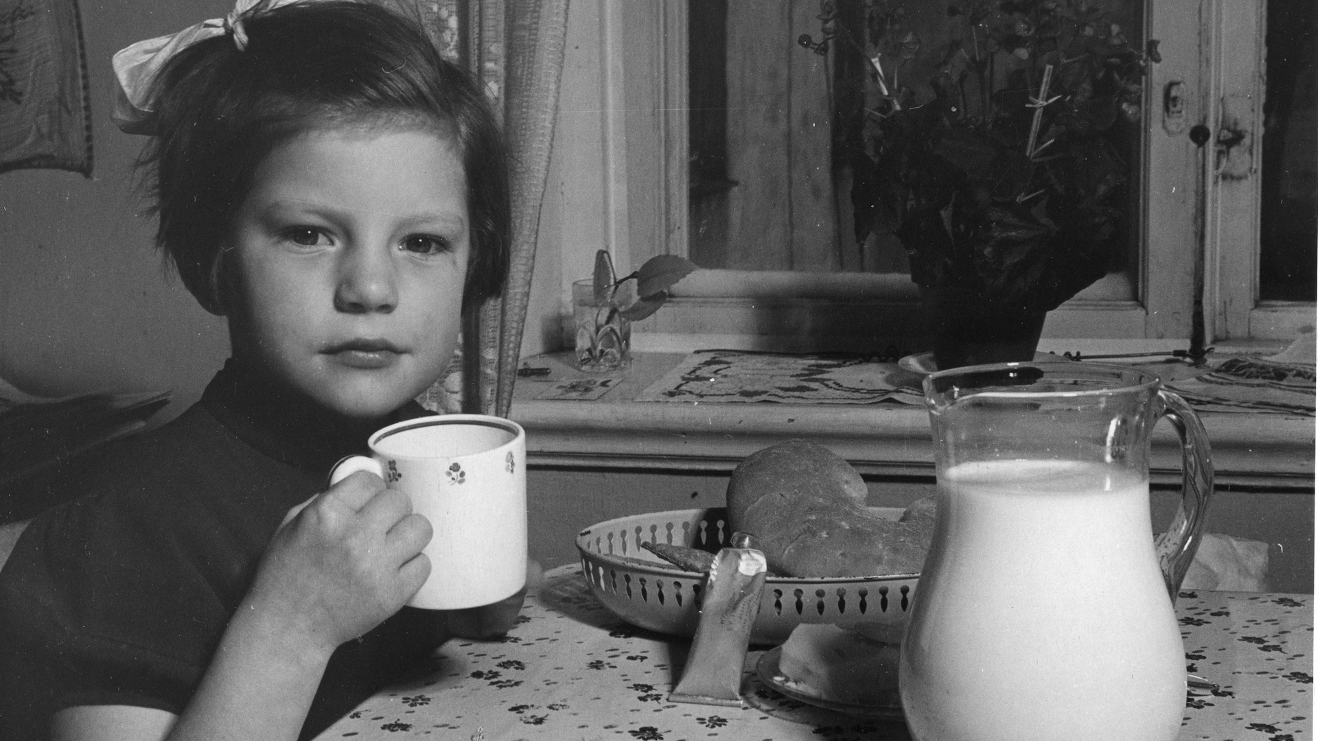 En flicka med en mugg, på bordet framför står en stor kanna mjölk. 