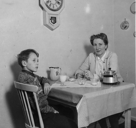 En pojke med ett glas mjölk, och en kvinna med en kaffekanna, vid ett köksbord.
