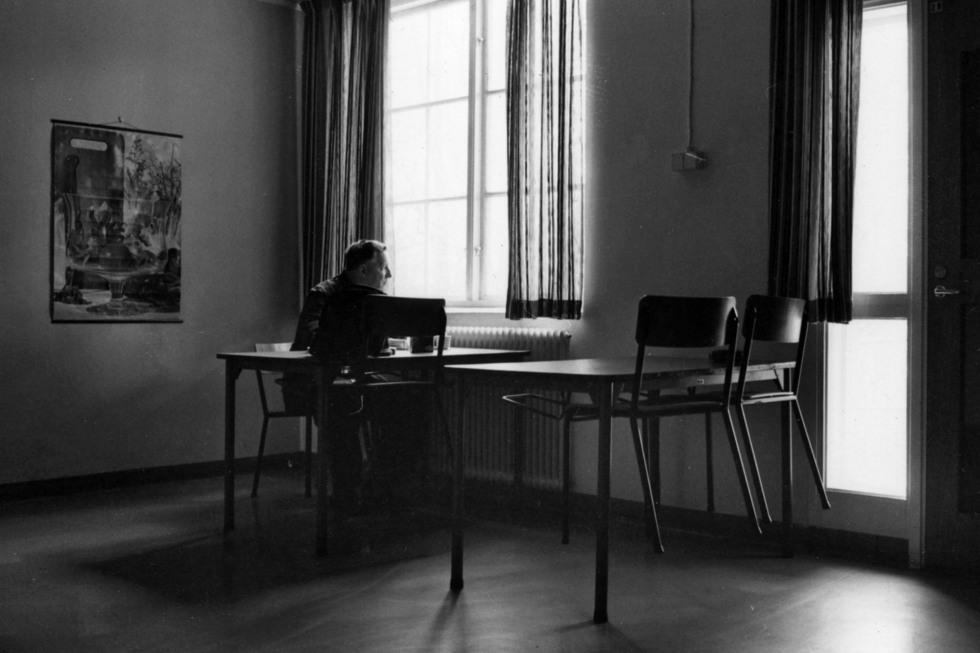 Ett svart-vitt foto på NK-verkstädernas lunchrummet. Stolarna har satts upp på bordet inför städning och en man tittar ur fönstret.