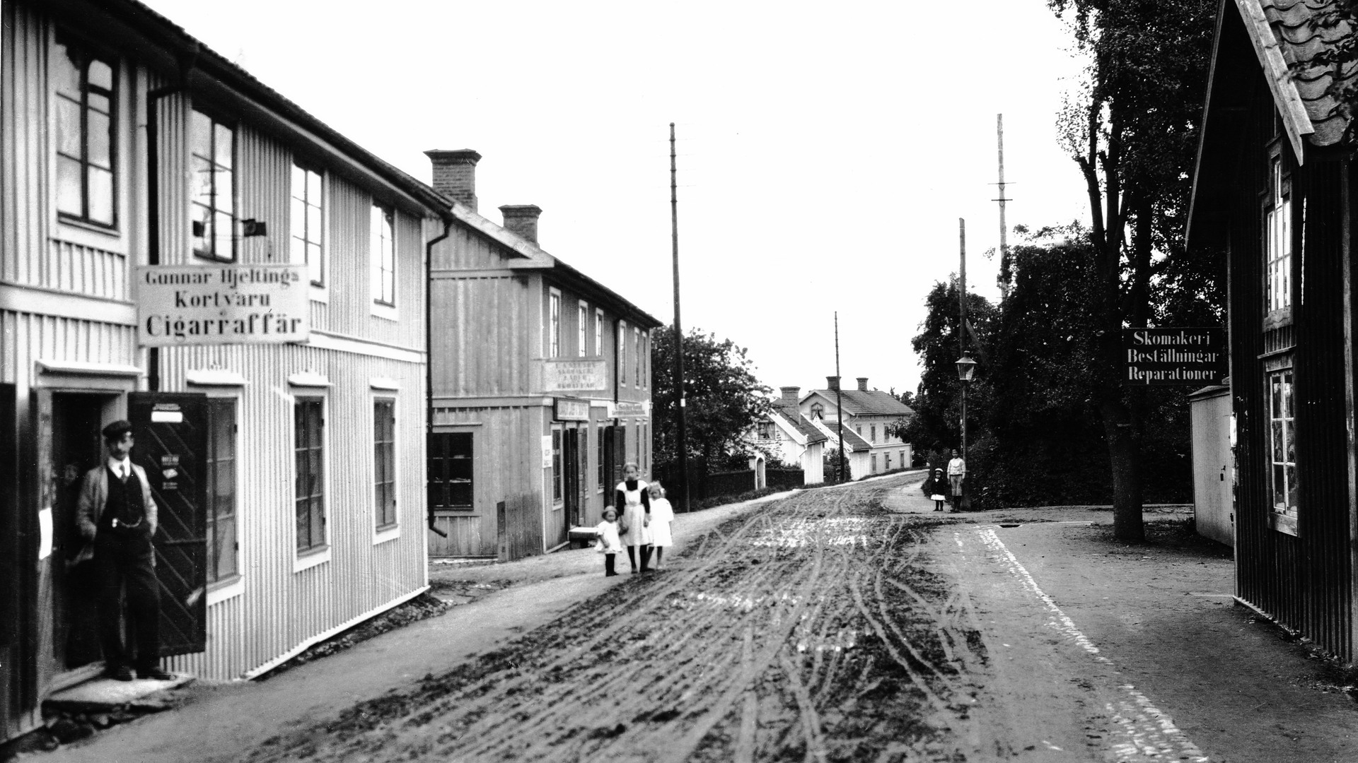 En gata med en cigarettaffär till vänster. Tre barn står i bakgrunden, en man på vänstra sidan.
