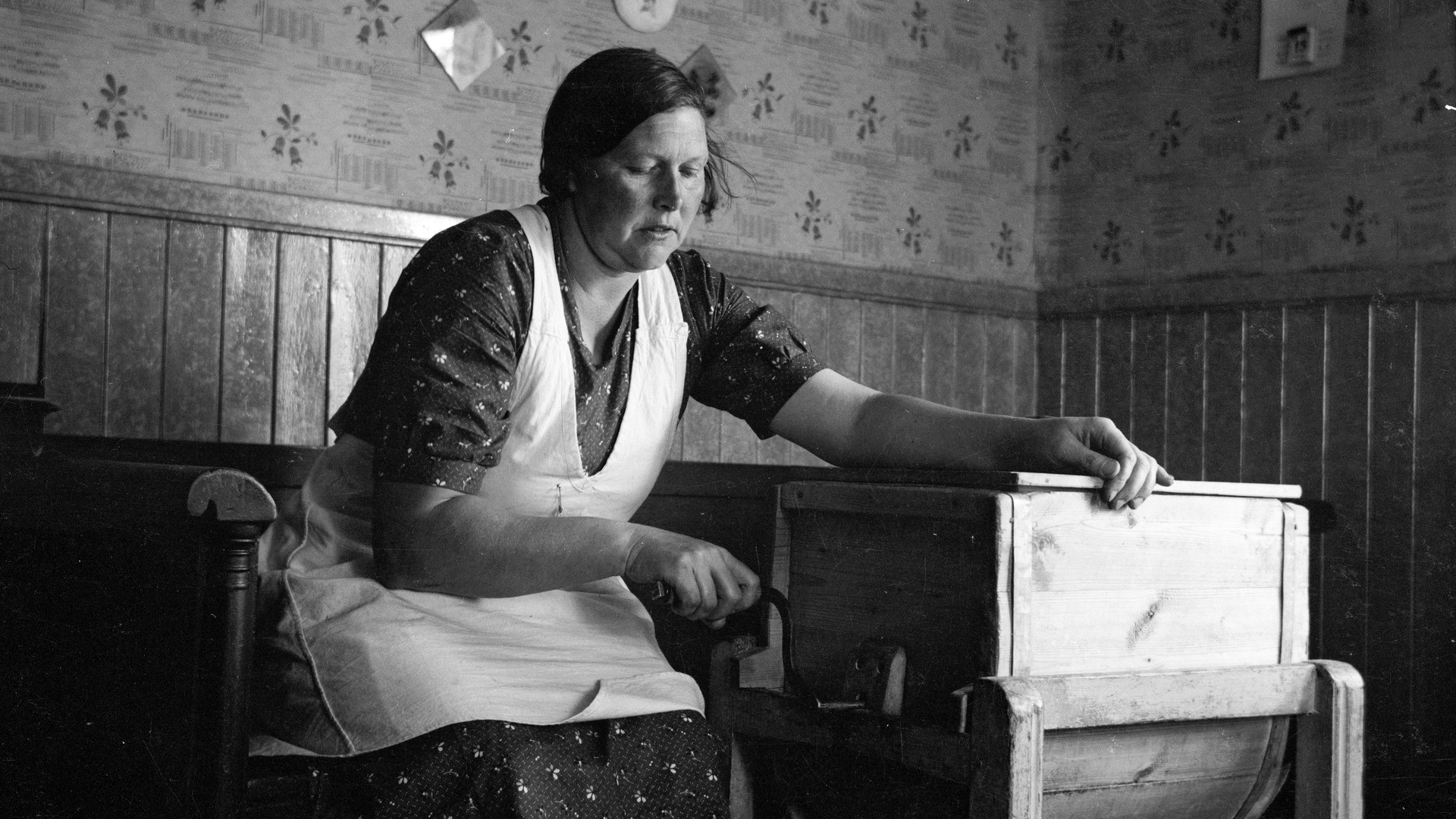 En kvinna med förkläde sitter ner och vevar på en stor smörkärna och kärnar smör.