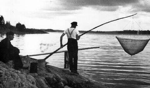 En man står på en klippa och fiskar med en stor håv. Bakom sitter en man och tittar på.